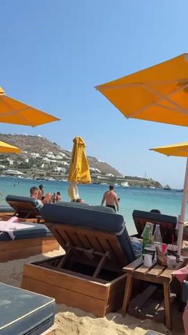 The video depicts a beach scene with several people lounging on beach chairs under bright yellow umbrellas. The water is a clear blue, and the sky is a bright blue with no clouds visible. In the background, there are hills with white buildings, suggesting a coastal town. Some people are in the water, and others are on the beach. There are also drinks and other items on small tables next to the beach chairs. The overall atmosphere appears relaxed and summery.