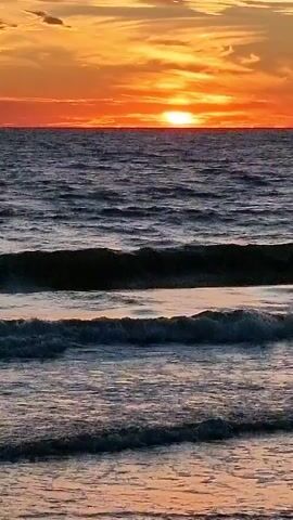 The video shows a close-up, low-angle shot of ocean waves crashing on a beach during sunset. The sun is setting over the horizon, casting a warm orange and yellow glow on the sky. The water is dark and choppy, with white foam cresting on the waves. The beach is made of sand and small pebbles. The video is shot in landscape orientation.