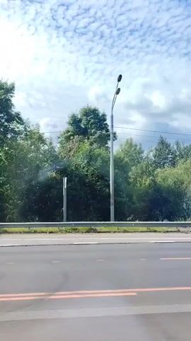 A video captures a roadside view of trees and a bright sky with scattered clouds. A white van drives past on the road.