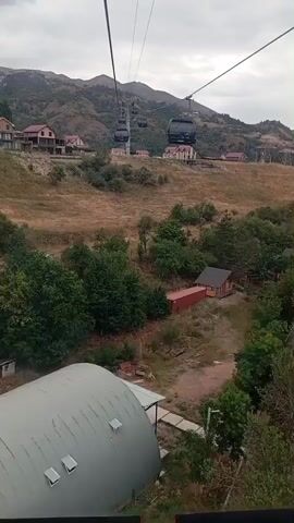 This is a low-angle, slightly shaky, first-person perspective video taken from inside a cable car moving along a cable line. The cable car is traveling uphill, and several other cable cars are visible ahead, also ascending. The background features a mountainous landscape with sparse vegetation, brown grass, and scattered trees. Several buildings, appearing to be houses and perhaps small structures, are situated on the hillside. In the foreground, a large, curved, silver metal roof structure, likely part of the cable car station, is visible. The sky is overcast.