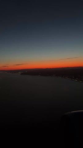 The video shows a sunset view from an airplane. The sky transitions from dark blue at the top to bright orange near the horizon. Below the horizon is a dark landmass with scattered lights indicating a city or town along the coast. The water is dark and reflects the colors of the sky. A portion of the airplane wing is visible in the lower right corner of the frame. A vapor trail is visible in the blue sky.