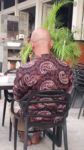The media file shows a rear view of a bald man wearing a patterned short-sleeved shirt and a sarong. He is sitting in a black plastic chair at a table, with another black chair stacked on top of another chair to his right. In the background, there is a building with a white sink mounted on a grey tiled wall, a mirror above it, and some potted plants with large green leaves. A person is visible walking in the background. The scene appears to be outdoors, possibly at a cafe or restaurant, with natural light suggesting it is daytime. There is no discernible text in the video.