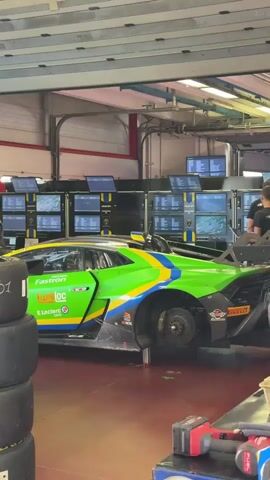 A race car pit lane with a green, blue, and yellow racing Lamborghini parked inside a garage. The car is partially disassembled, with its rear wheel missing and the chassis visible. Several people are working on or around the car, including one person in the foreground looking away from the camera, wearing a black t-shirt with a colorful striped logo. Another person in a black jacket and pants is leaning into the car from the side. A stack of tires is visible in the left foreground, with a number 6-01 written on the top tire. Numerous computer monitors displaying data are mounted on the wall behind the car. The garage has industrial lighting and a large overhead door in the background.