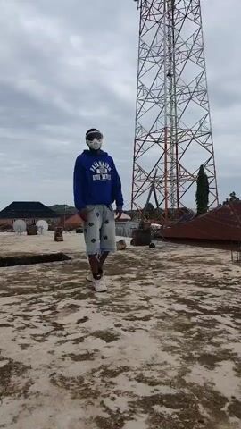 A person stands on a rooftop in front of a large tower. The person is wearing a blue hoodie, denim shorts, and goggles. The sky is cloudy. The ground is rough and appears to be made of concrete or stone. There are some buildings and vegetation in the background. The person is posing for the camera, with one leg extended.