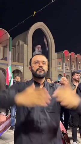 A man with a beard and glasses wears a black button-up shirt and tan pants. He stands with his arms raised and hands open, looking upwards. He is in front of a building with a large arched display featuring a portrait of an older man with a white beard and a black turban. Several other people are visible in the background. Iranian flags are present. The scene appears to be outdoors at night.
