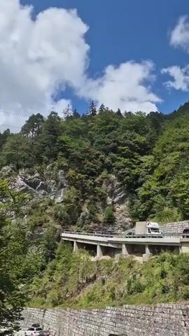 A truck drives on a road that curves along a mountain slope. Trees and rocks cover the mountainside, with a bright blue sky and white clouds overhead.