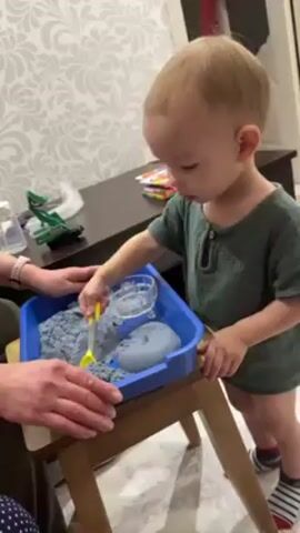 A toddler plays with gray kinetic sand in a blue tray on a small wooden table. The child is fair-skinned with blonde hair and is wearing a green short-sleeved shirt and striped socks. The child is using a yellow and white plastic spoon to scoop the sand. A clear plastic cup is also in the tray. A woman is sitting nearby, her hands resting on the tray. In the background is a dark brown table with various items on it, including a toy tractor and packages of colorful candies. The wallpaper is decorated with a gray and white paisley pattern.