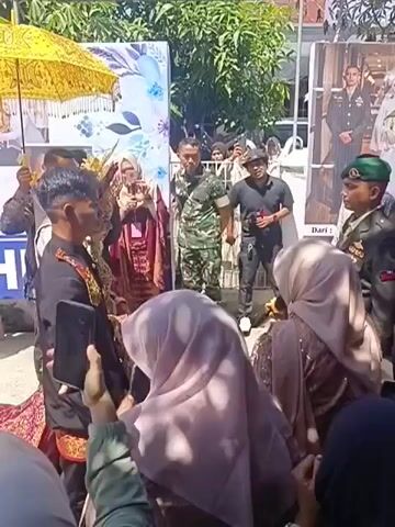 The video shows an outdoor wedding ceremony. A man and a woman are dressed in traditional wedding attire. The woman wears a red and gold ornate dress and a matching headpiece. The man wears a black outfit with gold embellishments and a tall, golden headpiece. They are standing facing each other, with a group of people gathered around them, many holding up phones to record the event.

A man in a military uniform is standing between the couple, facing them. To the right of the couple, another man in a military uniform, wearing a green beret, is also present. Behind the couple, there are two large posters. One poster features a floral design with blue and white flowers. The other poster displays a formal portrait of a couple in military and wedding attire.

The environment appears to be sunny, with trees visible in the background and a building with a red roof. Several individuals in the crowd are wearing hijabs. The overall atmosphere seems festive, with a focus on the wedding ceremony and the presence of military personnel, possibly indicating a connection to the military for one or both of the individuals being married.