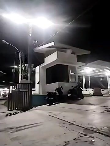 This video shows a nighttime scene in front of a small, modern-looking white building. Two motorcycles are parked in front of the building. One motorcycle is to the left of the building and the other is to the right. The building has a large dark-colored window or opening in the front. There are lights illuminating the scene, including a bright light from above. A fence is visible on the left side of the frame. The ground appears to be paved.