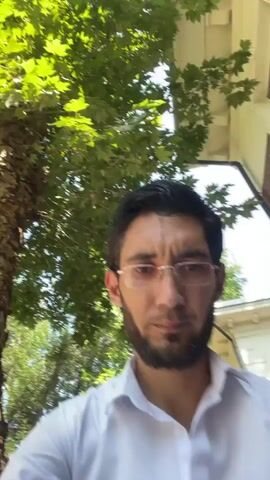 A man with a beard and glasses looks into the camera. He is wearing a white shirt. He appears to be outdoors with trees in the background. The camera is shaky, suggesting it is being held by hand.