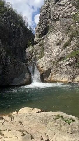 The video shows a scenic view of a waterfall cascading into a clear, turquoise-colored pool of water, surrounded by steep, rocky cliffs. The cliffs on both sides are a mixture of gray and white, with visible layers and crevices. Some sparse vegetation, including trees and bushes, cling to the cliff sides. The sky above is blue with scattered white clouds, suggesting a bright, partially sunny day. In the foreground, there are large, flat rocks leading up to the water's edge, forming a natural viewing platform. The waterfall itself is moderate in size, creating a frothy white splash as it hits the pool below. The scene evokes a sense of natural beauty and tranquility.