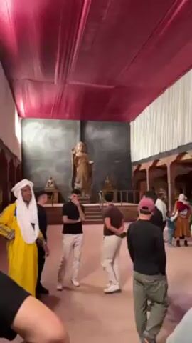 The video shows an indoor set with a red and white draped ceiling, set to resemble a Tibetan or Buddhist space. In the center of the room stands a tall statue, possibly of Buddha or a bodhisattva, placed against a dark backdrop. Several smaller statues are also visible. A group of people is gathered in the room, some conversing and others looking around. One person is wearing a yellow robe and a white turban. The camera moves to show the architectural details of the set, including decorated columns and a balcony. The overall impression is of a movie set or a themed environment designed to evoke a Tibetan or Buddhist aesthetic.