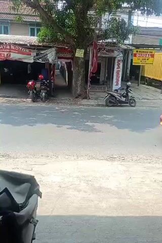 The video shows a street scene with a red van and several motorcycles. The van, a passenger van, is red and has its side door open. A person is visible in the driver's seat and another person is seated behind them. Several motorcycles are on the street, some in motion and some parked. One motorcycle, in the foreground, has a white helmet resting on its handlebars. The background consists of buildings with shop fronts and awnings, suggesting a commercial or market area. The street is paved, and there are sidewalks. The overall environment appears to be outdoors during the daytime, with sunlight casting shadows.