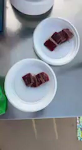 The video shows an overhead view of a metal tabletop with several small white plates. On most of the plates, there are dark red, rectangular pieces of food, possibly cured meat or fish like tuna, arranged in small stacks. One plate holds a pile of bright red, granular food, possibly a relish or sauce, with small sprinkles around it. Another longer, rectangular white plate also displays several pieces of the dark red food. A small clear container with a white powdery substance, likely salt, and part of a blue cutting board with a knife are also visible on the table. The scene appears to be an outdoor or semi-outdoor food preparation or serving area during the daytime, possibly at an event in Barbate, Spain, given the audio and regional context. Voices in the background are speaking Spanish, remarking on the food.