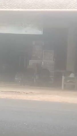 The video displays a hazy outdoor scene with a building in the background. In front of the building, there are two motorcycles parked side-by-side. A rack with several items is visible behind the motorcycles. To the right of the motorcycles, a chair is positioned outside. The ground appears dusty or sandy, and there is a road in the foreground, suggesting a potentially arid or dusty environment. The overall visibility is reduced due to the haze.