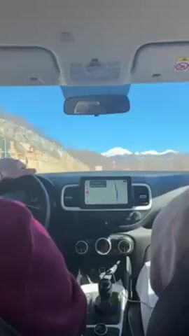The video shows the interior of a car from the perspective of the backseat. Two people are in the front, one driving and one looking at a phone. The car is driving on a road surrounded by mountains under a clear blue sky. The mountains in the distance are snow capped. The car's navigation system is displayed on the dashboard. The driver is wearing a burgundy shirt. The passenger is wearing a light brown shirt. The car has a manual transmission.
