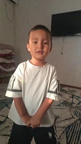 A young boy stands in front of the camera, wearing a white t-shirt with dark stripes on the sleeves and dark shorts. He is in a room with a tiled floor, a television mounted on the wall, and what appears to be an air conditioning unit above him. He smiles and makes faces at the camera.