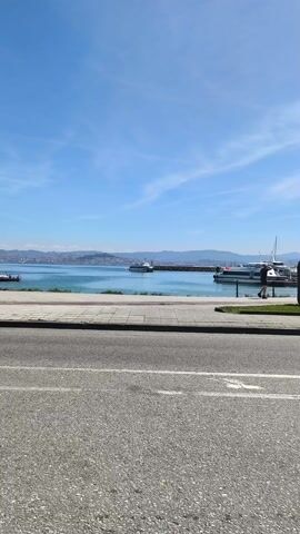 The camera steadily moves to the left along a gray asphalt road. A narrow concrete sidewalk separates the road from the waterfront. In the distance, beyond the turquoise water, lies a city on a hilly landscape under a clear, blue sky with faint white clouds. Several boats are visible on the water, some docked at a marina. A few people are walking along the waterfront in the distance. A lamppost stands on the sidewalk. A short row of trees is partially visible to the left of the lamppost.