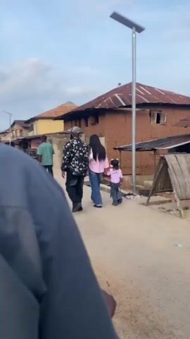 A video clip shows three individuals walking away from the camera on an unpaved dirt road. The leftmost person is an adult wearing a black and white patterned long-sleeved top and dark pants. The middle person is an adult female with long dark hair, wearing a light pink long-sleeved shirt, blue jeans, and carrying a white shoulder bag; she holds the hand of a young child to her right. The child wears a light purple long-sleeved top and dark pants. On the right side of the road, a tall, grey metallic pole with a solar panel and a light fixture stands upright. Behind the walking figures is a large, rustic building constructed of reddish-brown material, possibly mud or clay, topped with a rusty, corrugated metal roof. The roof has visible undulations and some discoloration. Small, dark window openings are visible on the building's facade. A wooden A-frame structure is partially visible on the far right. The sky is bright and overcast.