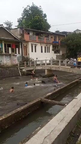 The video shows children swimming and playing in a river that runs through a residential area. The river has a moderate current and contains some large rocks. The children are of various ages and are wearing swimsuits. They are splashing, throwing a blue ball, and swimming in the water. On either side of the river are concrete banks and drainage structures. A small bridge spans the river, and people are standing on it observing the children. Houses and trees are visible in the background. The sky appears overcast, suggesting a cloudy day.