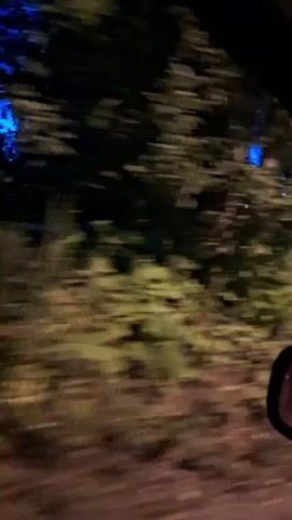 The video is a blurry, first-person perspective shot from inside a vehicle, likely taken at dusk or night. The camera is moving, causing motion blur, and the view is primarily of trees and vegetation on the left side of the road, with some houses and a clear blue sky visible in the distance on the right. The lighting suggests twilight or nighttime. The scene appears to be a rural or suburban area with a dirt or gravel road. There are no people or specific actions clearly depicted due to the blur and low light.