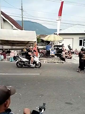 The video shows a street scene, possibly during a parade or cultural event. A person in the foreground, wearing a black outfit and a backpack, holds a long stick. In the background, a group of people in traditional attire, some with elaborate costumes and headdresses, are gathered. Two red and white flags are visible, suggesting a national or regional celebration. There are also several vehicles, including motorcycles and a small blue truck carrying speakers. People are seated along the side of the road, observing the event. The environment appears to be a town or village, with buildings and mountains in the distance. The overall atmosphere seems festive.