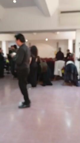 A group of people are gathered in a large room with a light-colored floor and ceiling. The room appears to be a hall or an event space, with chairs and tables visible in the background, some covered with dark fabric. Many of the individuals are wearing dark clothing, possibly black, suggesting a uniform or a specific dress code for an event. In the foreground, a person wearing a black long-sleeved shirt and black pants stands facing away from the camera, holding a white object in their hands. Other individuals are dispersed throughout the room, some standing and talking, while others appear to be seated or preparing to be seated. The lighting is bright, casting soft shadows on the floor. The overall atmosphere seems to be one of anticipation or waiting.