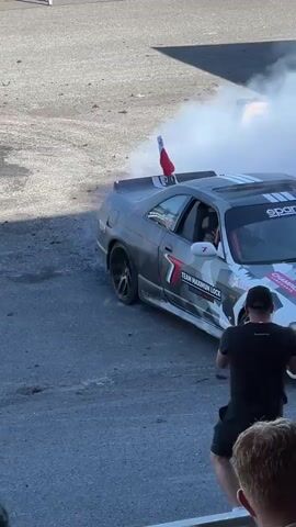 A drift car is parked on asphalt, emitting smoke. A person in a helmet and pink racing suit is on a motorcycle next to it, with one hand raised. Another person in a helmet and a light-colored shirt is on a dirt bike to the left of the car. Two people are standing in the foreground, watching the scene. The background features a green grassy area with a fence and some poles. The car has racing decals and a Polish flag attached to its spoiler.
