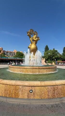 A golden statue of a cartoon character is shown in a fountain in front of an entrance to a theme park. The character is holding up a "50" with stars around it, indicating an anniversary celebration. The fountain has multiple jets of water spraying upwards, and the water is a greenish color. The background shows a rocky structure with a waterfall, and a building with an entrance and several people in line. The sky is clear and blue, and there are trees and other foliage visible. The overall impression is a bright, sunny day at a popular tourist destination.