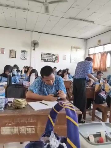A classroom scene viewed from the front, with rows of students seated at wooden desks. The students, mostly young women wearing hijabs and blue uniforms, are facing forward and appear to be engaged in some activity, possibly writing or studying. In the foreground, a man in a blue patterned shirt is seated at a desk, looking down at a small black object, possibly a mobile phone. To the right, a teacher, also in a blue patterned shirt, stands and interacts with a student. The classroom is brightly lit, with fluorescent lights on the ceiling and natural light coming from windows on the left. Various items are visible on the desks, including books, water bottles, and stationery. The overall atmosphere appears to be one of a typical school day.