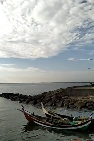 The video shows a tranquil view of a body of water, likely a bay or harbor, with two colorful fishing boats docked near a rocky breakwater. The boats, one with red and the other with yellow accents, are small and appear to be traditional in design. Fishing nets and gear are visible inside one of the boats.  The breakwater is constructed from large, dark rocks, and a concrete walkway extends from it. In the background, the water stretches to the horizon under a sky filled with fluffy, white clouds. The overall atmosphere is calm and serene.