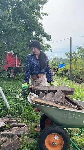 A young woman with long dark hair is working in a backyard. She is wearing a blue patterned shirt, tan shorts, and green gardening gloves. The woman is bending over and picking up pieces of wood. She places the wood into a green wheelbarrow that is already filled with more pieces of wood. The wheelbarrow has two orange-rimmed wheels. In the background, there is a red building, trees, and a fence. The ground is covered in grass and scattered pieces of wood. The sky is cloudy.