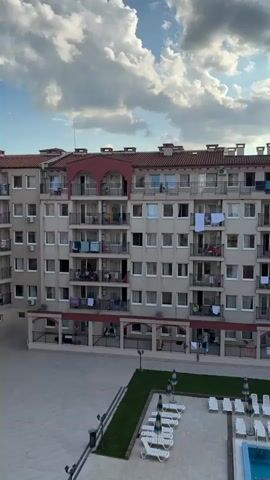 The video pans across a resort complex. In the foreground, a long blue swimming pool with lane markers is visible, bordered by rows of white lounge chairs and blue and yellow striped umbrellas. Beyond the pool is a meticulously maintained green lawn. In the middle ground, a multi-story building with numerous balconies is seen. Laundry is hanging on some of the balconies. Another taller building with a distinctive turret-like design stands to the right of the first building. The sky above is filled with dramatic, cloud-filled formations, with the sun peeking through in the upper right corner. The overall scene suggests a sunny day at a vacation destination.