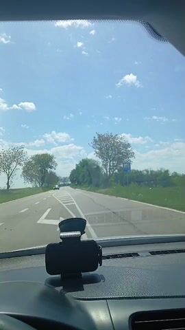 The video shows a car driving down a road on a sunny day. The sky is blue with scattered white clouds. The road is straight and has white lines marking the lanes and pedestrian crossings. There are trees on either side of the road, and a sign is visible on the right side. The camera is mounted on the dashboard of the car. Music is playing in the background.
