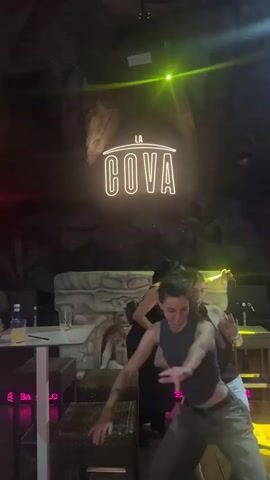 The video shows a dimly lit indoor space with a neon sign that reads "LA COVA." Several young women are visible, some dancing and interacting with each other. One woman in the foreground is wearing a grey tank top and olive green pants and is in motion. To her left, two women in black dresses are standing and talking. Another woman in a white dress is visible in the lower right corner, laughing. The background appears to be a textured, cave-like wall. There are tables and chairs in the scene, suggesting a social gathering or a nightclub.