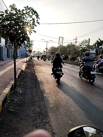 A first-person perspective video shows a person riding a motorcycle on a street lined with trees and buildings on the left. The street is paved and appears to be in a town or city. There are several other motorcycles on the road, some of which are overtaking the camera. The sun is shining brightly, casting long shadows. The time of day appears to be late afternoon or early evening. The video is short and shows a continuous action of riding a motorcycle.
