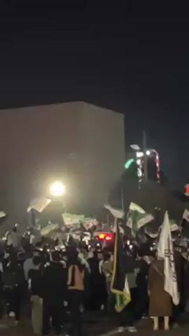A crowd of people gather at night, many waving flags, seemingly in celebration or protest. The flags appear to be mainly white and green, with some bearing stars and crescents.  The group is densely packed, and the environment includes streetlights, buildings, and a structure with colorful lights.  The atmosphere appears lively and energetic. The camera angle looks up toward the crowd and a street lamp.