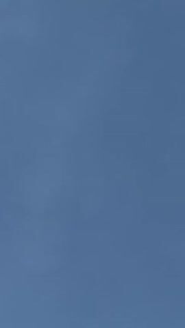 A single airplane is flying across a clear, blue sky. The plane is dark in silhouette against the bright sky, and it appears to be at a high altitude, moving from left to right across the frame. No clouds are visible. The setting is an open sky, likely during the day, above Gangloff, Germany. No people or text are visible.