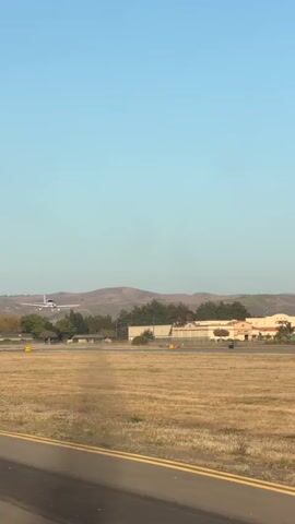 A small white airplane is seen from the side as it ascends into a clear blue sky. The plane is in the left half of the frame, and the background consists of rolling hills and buildings. The foreground is a dry, grassy field with a paved runway. The sun appears to be low in the sky, casting long shadows.