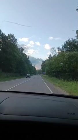 The video is shot from the interior of a car as it drives down a two-lane road surrounded by dense forest. The sky is partly cloudy with hints of sunset. A dark-colored vehicle is seen driving in the opposite lane. Road signs are visible on the right side of the road, including a triangular warning sign. The interior of the car, including the dashboard, is visible in the foreground.