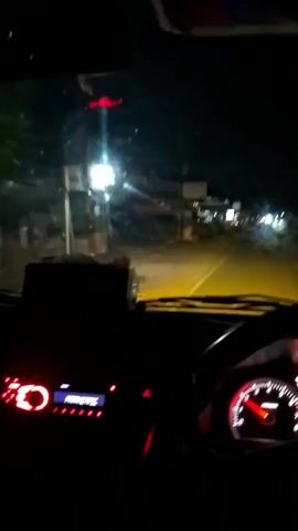 This is a first-person perspective video from inside a vehicle at night, likely in Tambonbaroh, Indonesia. The driver's dashboard is prominently featured, with illuminated gauges showing the speedometer and tachometer. The headlights cast a yellowish glow on the road ahead, illuminating a rural or semi-rural setting. On either side of the road, there are glimpses of buildings and trees, some with lights on, suggesting a small village or roadside businesses. The overall mood is one of travel or commuting in the darkness. The time of day is clearly night due to the absence of natural light and the illumination provided by the car's headlights. There is no visible text in the video.