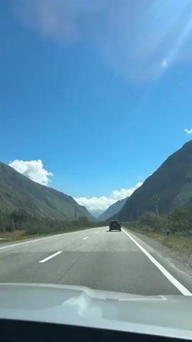 This is a first-person perspective video, likely from inside a vehicle. The scene shows a car driving on a paved road that winds through a mountain valley. The sky is bright blue with a few scattered white clouds, and the sun is casting lens flares. On either side of the road are steep, green mountains. There is another dark-colored vehicle ahead, also traveling in the same direction. Power lines and poles are visible along the right side of the road. The road has white lane markings.