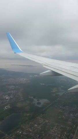 The video shows a view from an airplane window. We see a wing of the plane with a blue tip. Below, there's a landscape consisting of buildings, trees, and bodies of water like ponds and a larger body of water in the distance. The sky is overcast with grey clouds.
