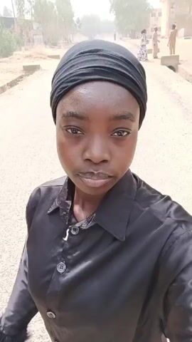 The video shows a young, dark-skinned person with a black head covering and a black button-down shirt. They are standing outdoors on what appears to be a street or road with trees and buildings in the background. The person holds a dark, feathery object behind their head, resting their cheek on their hand. They are looking directly at the camera with a neutral expression. The scene seems to be recorded during the daytime with slightly hazy conditions. Other people can be seen walking in the background.