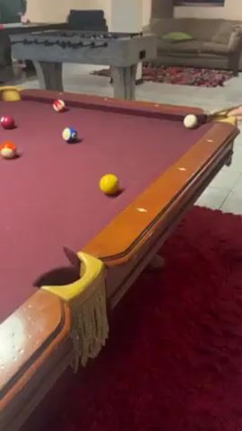 The scene captures a recreational space with a pool table as the central focus. The pool table has a deep burgundy felt surface and is surrounded by a red shag rug. Several pool balls, including a yellow one, are scattered across the table. One of the pool balls is struck, causing the other balls to scatter, and the black ball falls into the pocket. In the background, a foosball table is visible, with a person standing near it. There is also a tan-colored sofa with a green cushion, suggesting a comfortable lounge area within the room. The room is well-lit, highlighting the various gaming and relaxation features.