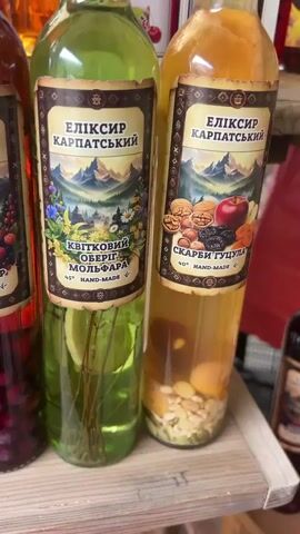 The video shows a close-up, panning shot of several bottles filled with various liquids and ingredients. The bottles are standing upright on a wooden surface. Each bottle has a label with a mountain landscape and Cyrillic text. From left to right, the bottles appear to contain: dark red liquid with whole berries, light green liquid with herbs and a lemon slice, amber liquid with dried fruit and nuts, light yellow liquid with citrus slices, dark red liquid with whole berries, and another amber liquid with dried fruit and nuts. The overall impression is a display of artisanal beverages or infusions.