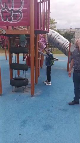 The video shows a playground with a large red and brown climbing structure. Three children are visible on the structure, two climbing and one at the top. A man in a blue patterned shirt and jeans stands to the right of the structure, holding his hand to his forehead. Another child, wearing a black and white checkered hat and a blue jacket with a backpack, is climbing on the side of the structure. The ground is a blue rubber surface. A silver tube slide curves away from the structure. The background shows trees and a building.