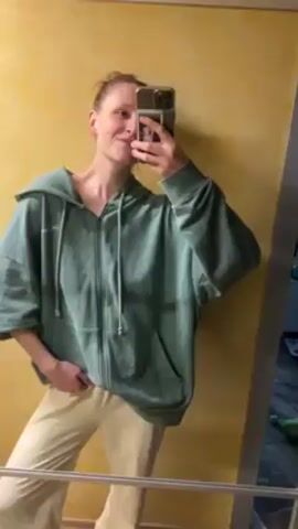 A slender young adult is standing in front of a bathroom mirror, recording a selfie video with an iPhone. She is wearing a light green, oversized zip-up hoodie and beige pants. The background is a pale yellow wall. Initially, she poses casually with one hand in her pocket. She then makes a peace sign with her free hand. Finally, she briefly steps to the side, and the camera focuses on the yellow wall.