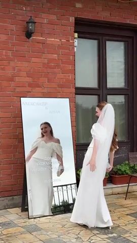 A bride in a white wedding dress and veil stands facing a large mirror. Her reflection is visible in the mirror. The mirror has text inscribed on it, which includes names, a greeting, and a date. The bride raises her veil and appears to be posing for the camera. The background consists of a brick wall and a door.