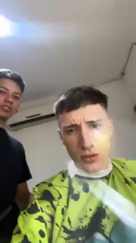 The video shows a young man in a barber chair getting a haircut. He is wearing a bright green haircutting cape with a colorful design. The barber, wearing a black shirt, stands behind him and trims the man's hair. The man looks into the camera. The scene is set inside a barber shop. An air conditioner is visible on the wall. The barber makes a gesture toward the camera.